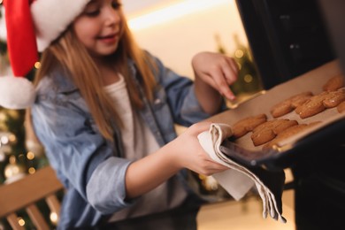 Photo of Little child in Santa hat taking baking sheet with Christmas cookies out of oven indoors, focus on hand