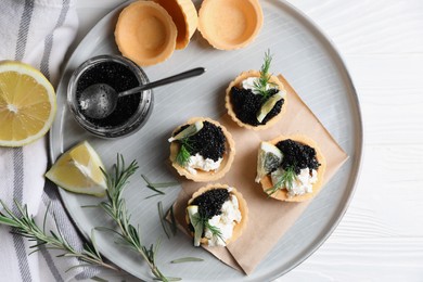 Photo of Plate of delicious tartlets with black caviar, cream cheese and lemon served on white wooden table, top view