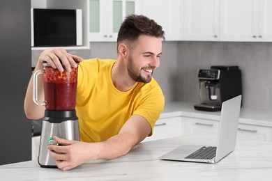 Photo of Happy man making smoothie while watching cooking online course in kitchen. Time for hobby