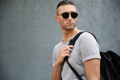 Handsome young man in stylish sunglasses with backpack near grey wall, space for text