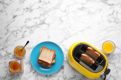 Yellow toaster with roasted bread, glass of juice and jam on white marble table, flat lay. Space for text
