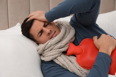 Photo of Ill man with hot water bottle on bed suffering from cold at home