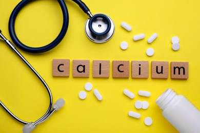 Word Calcium made of wooden cubes with letters, stethoscope and pills on yellow background, flat lay