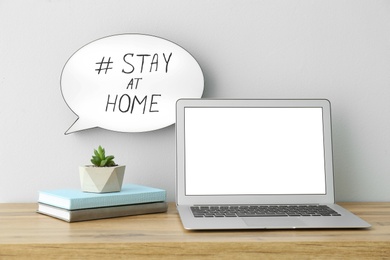 Laptop, books, houseplant and speech bubble with hashtag STAY AT HOME on white wall. Message to promote self-isolation during COVID‑19 pandemic