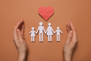 Woman protecting paper family figures and red heart on brown background, top view. Insurance concept
