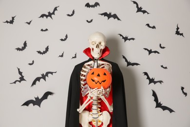 Skeleton in cloak with pumpkin and paper bats on light wall