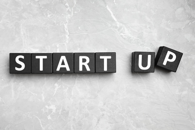 Words START UP made with black cubes on light grey marble background, flat lay
