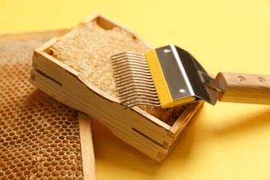 Hive frames with honeycombs and uncapping fork on yellow background, closeup. Beekeeping