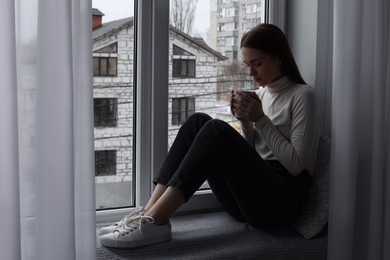 Melancholic young woman with cup of drink near window indoors, space for text. Loneliness concept