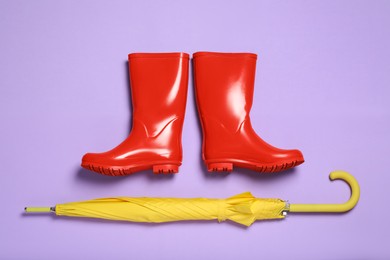 Pair of red rubber boots with yellow umbrella on violet background, flat lay