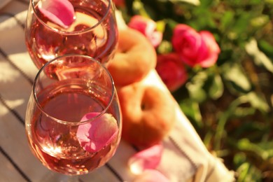Photo of Glasses of delicious rose wine with petals on white picnic blanket outside, closeup. Space for text