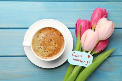 Cup of aromatic coffee, beautiful pink tulips and Good Morning note on light blue wooden table, flat lay