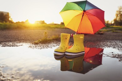 Yellow rubber boots and umbrella in puddle outdoors, space for text. Autumn walk
