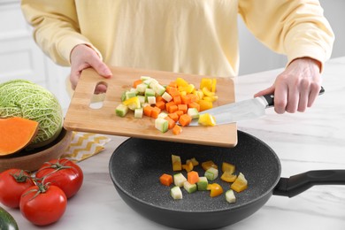 Photo of Woman pouring mix of cut vegetables into frying pan at table in kitchen, closeup