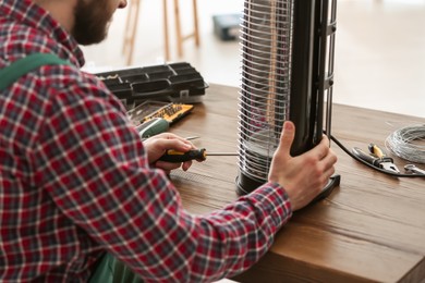 Professional technician repairing electric halogen heater with screwdriver at table indoors, closeup