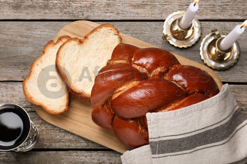 Cut homemade braided bread, goblet and candles on wooden table, flat lay. Traditional Shabbat challah