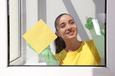 Photo of Happy young woman cleaning window glass with sponge cloth and spray indoors