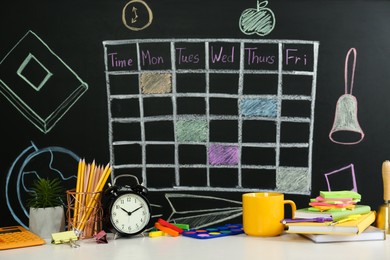 Different stationery, alarm clock and cup on white table near blackboard with drawn school timetable
