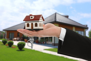 Real estate agent holding key and house model against beautiful villa, closeup