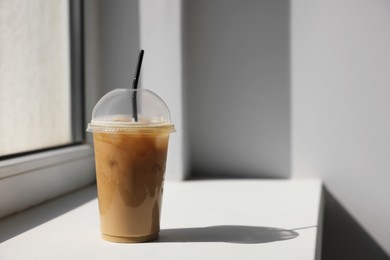 Photo of Plastic takeaway cup of delicious iced coffee on window sill indoors, space for text