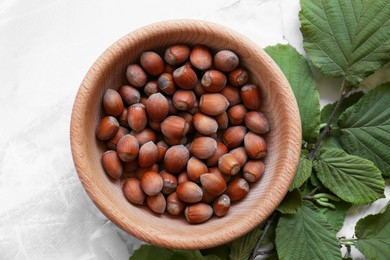 Photo of Tasty hazelnuts in wooden bowl and branch on light marble table, flat lay