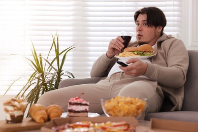 Overweight man with plate of burgers and French fries watching TV on sofa at home