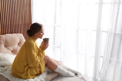 Beautiful young woman with cup of drink near window at home, space for text. Winter atmosphere
