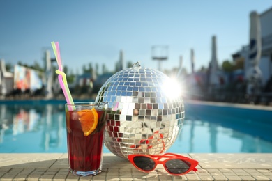 Shiny disco ball, refreshing cocktail and sunglasses on edge of swimming pool. Party items