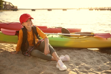 Happy girl sitting near kayak on river shore. Summer camp activity