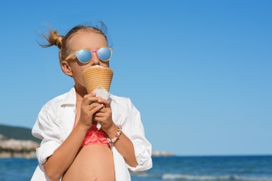 Adorable little girl eating delicious ice cream near sea on sunny summer day, space for text
