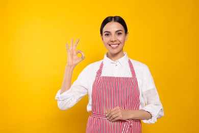Photo of Young woman in red striped apron showing okay gesture on yellow background