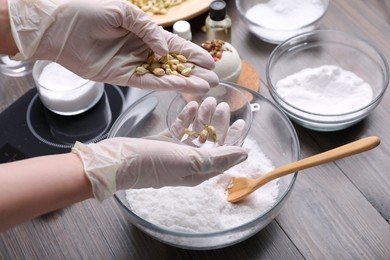 Woman in gloves filling bath bomb mold with dried flower buds at wooden table, closeup