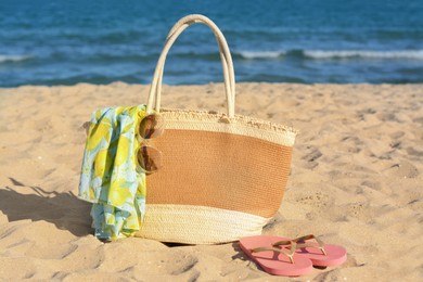 Photo of Straw bag with beach wrap, sunglasses and flip flops on sandy seashore. Summer accessories