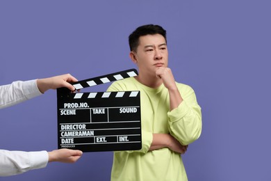 Photo of Asian actor performing while second assistant camera holding clapperboard on purple background, selective focus. Film industry