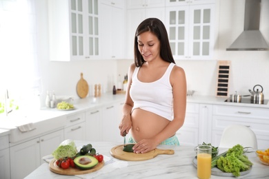 Young pregnant woman cutting avocado at table in kitchen. Taking care of baby health
