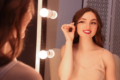 Photo of Beautiful young woman in elegant dress applying mascara near mirror indoors