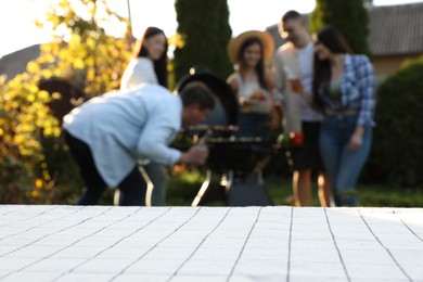 Empty white wooden table and blurred view of friends having barbecue party outdoors. Space for text
