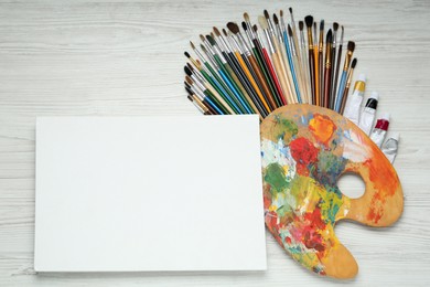 Blank canvas, brushes, palette and paints on white wooden table, flat lay. Space for text