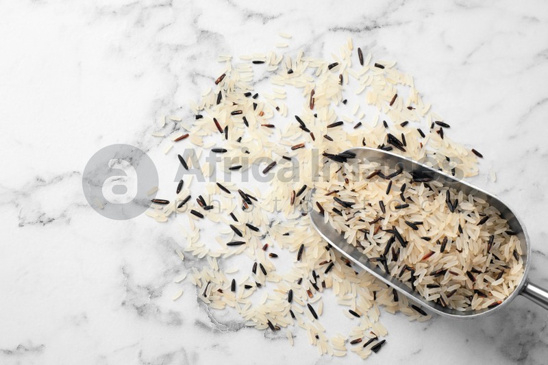 Mix of brown and polished rice with scoop on white marble table, top view