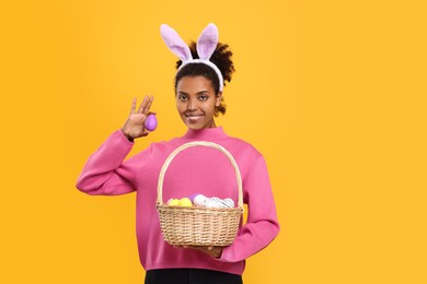 Photo of Happy African American woman in bunny ears headband holding wicker basket with Easter eggs on orange background. Space for text