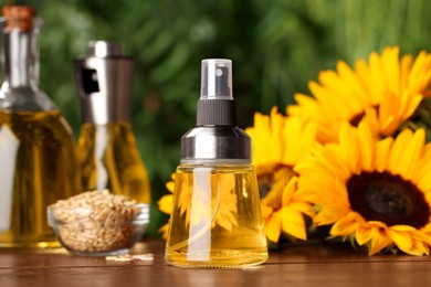 Spray bottle with cooking oil near sunflower seeds and flowers on wooden table against blurred green background