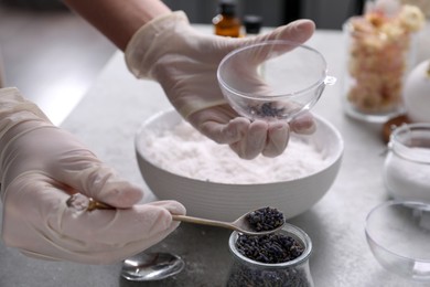 Woman in gloves filling bath bomb mold with dried flower buds at grey table, closeup