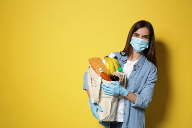 Female volunteer in protective mask and gloves with products on yellow background, space for text. Aid during coronavirus quarantine
