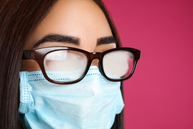 Young woman with foggy glasses caused by wearing disposable mask on pink background, closeup. Protective measure during coronavirus pandemic