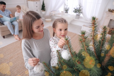Mother with daughter decorating Christmas tree together while father and son sitting on sofa at home