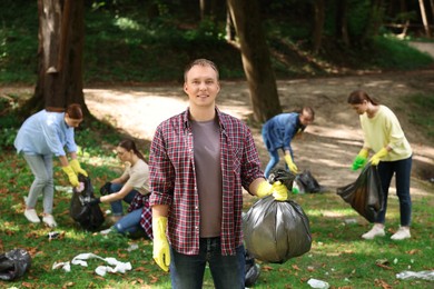 Photo of Young man with plastic bag and group of people collecting garbage in park