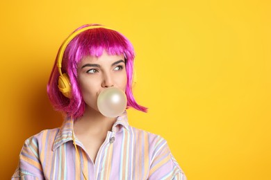 Fashionable young woman in colorful wig with headphones blowing bubblegum on yellow background, space for text