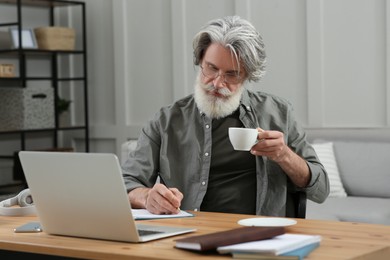 Middle aged man with laptop, notebook and cup of drink learning at table indoors
