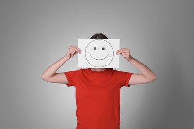 Photo of Woman hiding behind sheet of paper with happy face on grey background