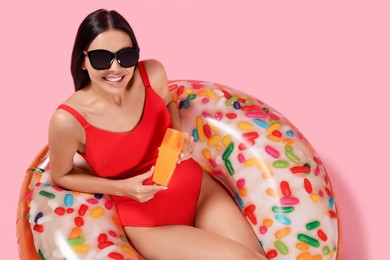 Young woman with sun protection cream on inflatable ring against pink background, above view
